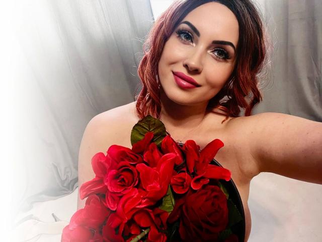 MystischLea69-hot with red roses, holding bouquet against white background, smiling with pink lipstick, wavy red-brown hair, and dark eyebrows. Boobs covered by roses. Soft lighting.