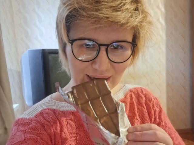SabrinaShine with short blonde hair and glasses, eating a chocolate bar, holding it with her right hand, in a pink knit sweater, looking slightly shy.