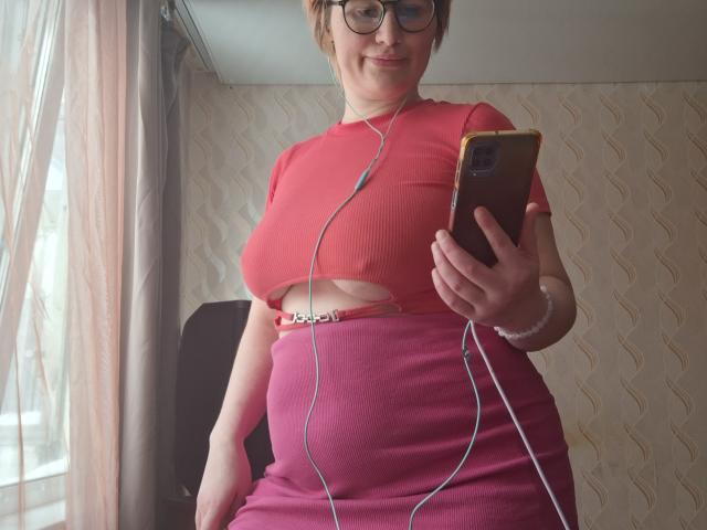 SabrinaShine with glasses, pink ribbed crop top showing her tits, pink high-waist skirt, white earbuds, holding phone, smiling, short brown hair, standing by window.