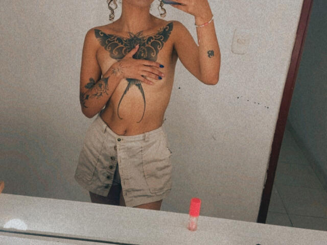 SilvanaXOwen with topless, tattooed chest, hand covering small tits, white button-up skirt, curly hair, holding phone, mirror selfie, white wall, red bobby pin on countertop.