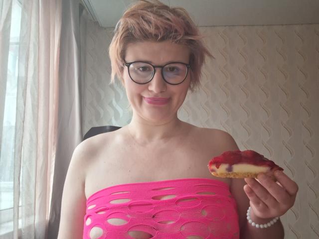 SabrinaShine with short pink hair and glasses, wearing a pink, cut-out, strapless top, holding a donut with red filling, smiling, showing her tits, and wearing a white bracelet.