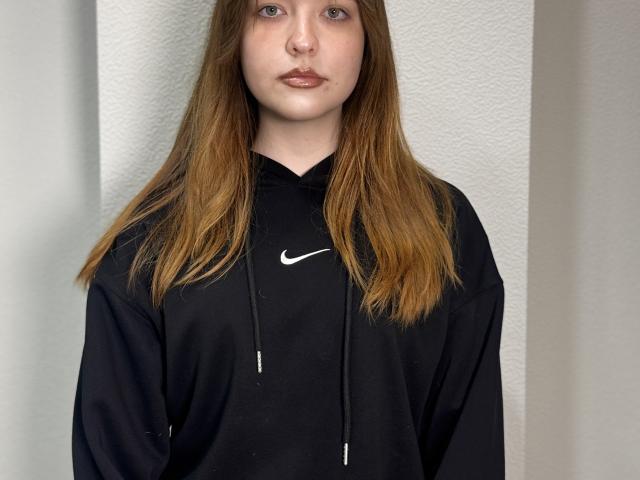 LilianDolfi with long brown hair, wearing a black Nike hoodie, looking straight at the camera with a neutral expression, soft light on her pale skin, subtle pink lipstick.