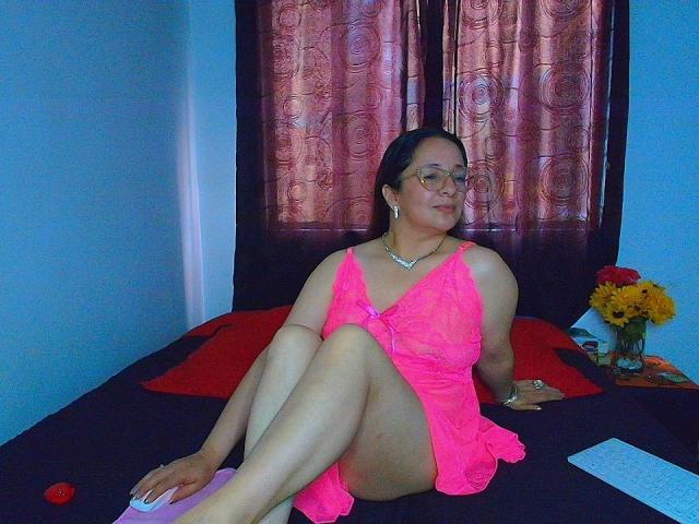 VictoriaRousse, chubby Latina woman with glasses, pink lingerie, bare ass, and small tits, sitting on black bed, legs crossed, flower vase on right.