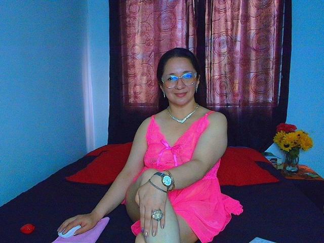 VictoriaRousse, wearing a neon pink dress, glasses, and a watch, sitting on a black bed with red pillows, has a cute ass and a small, cute pussy.