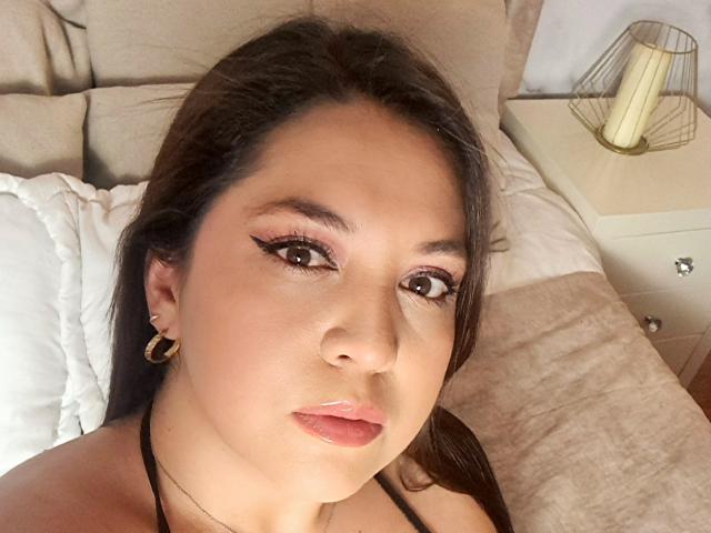 Paula, com olhos castanhos, cabelos longos e escuros, e batom rosa, usando brincos de argola dourados, um top preto de alças e maquiagem discreta, deitada na cama com um travesseiro branco..