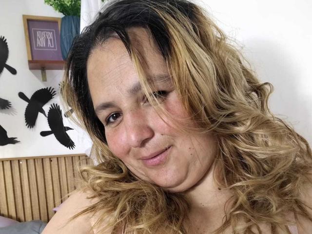 RouseWright has wavy, blonde-to-brown ombre hair, light skin, and a slight smile, with a small mole above her right lip. She's wearing a pink top, looking cute.