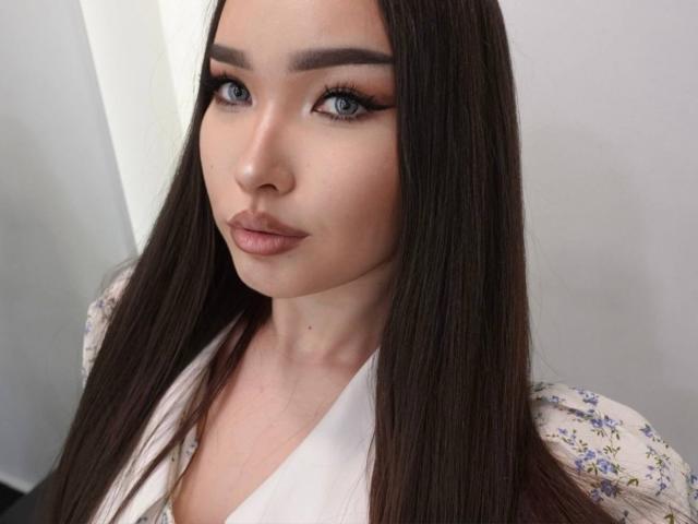 RebecaPfendler has long brown hair, blue eyes, and full pink lips with subtle makeup. She wears a floral-patterned shirt with a white V-neck.