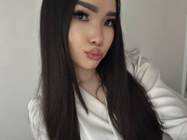 RebecaPfendler with long dark hair, thick eyebrows, and pink lips, wearing a white shirt, looking at the camera with a pouty expression. Her tits are not visible.