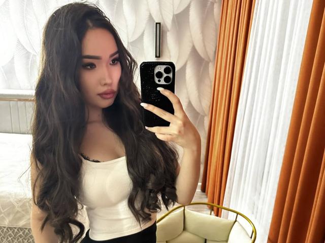 Rebeca Pfendler mit langem, gewelltem, dunklem Haar, trägt ein weißes Tanktop, hält ein Telefon, macht ein Selfie im Spiegel und zeigt ihre Brüste und einen kleinen schwarzen String..
