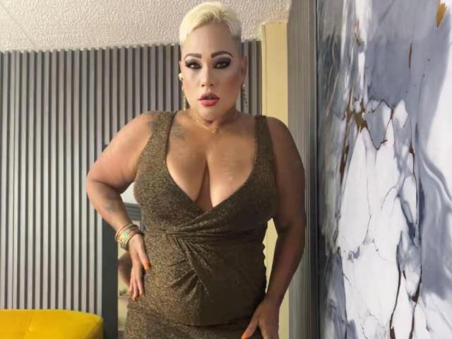 GianellaCox with blonde hair, large tits, and a curvy ass, wearing a deep brown, sparkly dress, standing with hands on hips, looking at the camera.