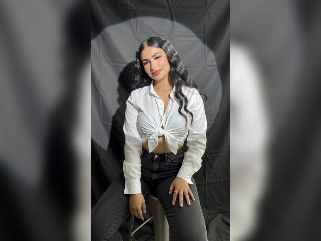 MiilaWx with long black wavy hair, white button-up shirt tied below breasts, black high-waisted jeans, sitting, smiling, hands on knees, soft light, neutral expression, subtle makeup.