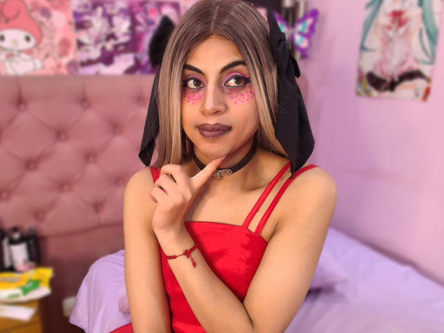 EimyFlorian with blonde hair, pink eye makeup, black headpiece, red dress, collar, finger near mouth, looking at camera, pink bed in background. Boobs not visible. Pussy not visible.