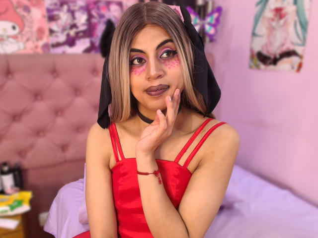 EimyFlorian with pink eyeshadow, red lipstick, and a red dress, touching her chin, wearing a black headpiece, in a pink bedroom. Blonde hair, light skin, cute face.