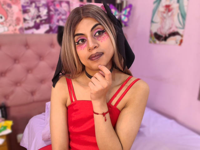 EimyFlorian with pink face paint, red dress, black headband, finger to mouth, choker, pink bed, purple wall, anime posters, and butterfly wall decoration. Blonde hair, brown eyes, looking cute.