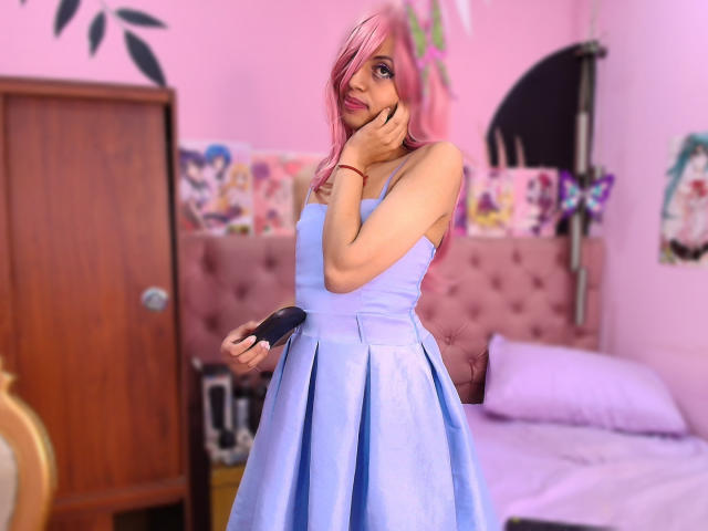 EimyFlorian with pink hair, wearing a blue strapless dress, holding a black brush, hand on chin, smiling, in a pink-themed bedroom. Boobs are small, ass is not visible.