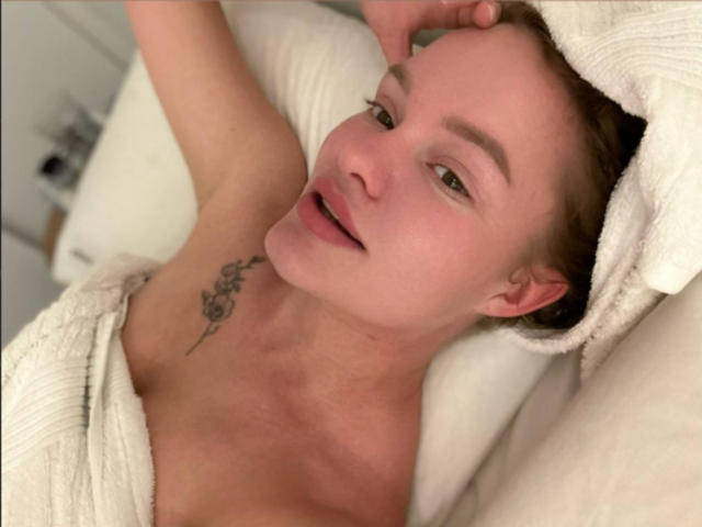 MySchellenger, sdraiato a letto, ha un asciugamano bianco avvolto attorno alle spalle nude, che lascia intravedere un piccolo tatuaggio sulla parte superiore del petto, con un'espressione rilassata e leggermente a bocca aperta..