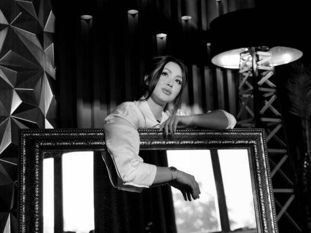 Black and white photo of AlyceSkagen leaning on a large framed mirror, wearing a white shirt, gloves, and looking off to the side with a sultry expression.