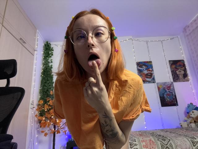 SarahRousen with orange hair, glasses, and colorful hair clips, tongue out, finger in mouth, wearing an orange shirt, tattoo on right arm, in a brightly lit room.