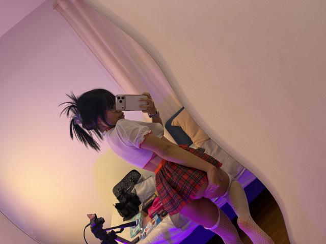 VannesaSaenz with a ponytail, wearing a white crop top, red plaid skirt, and white knee socks, taking a mirror selfie, showing her ass, in a dimly lit room.