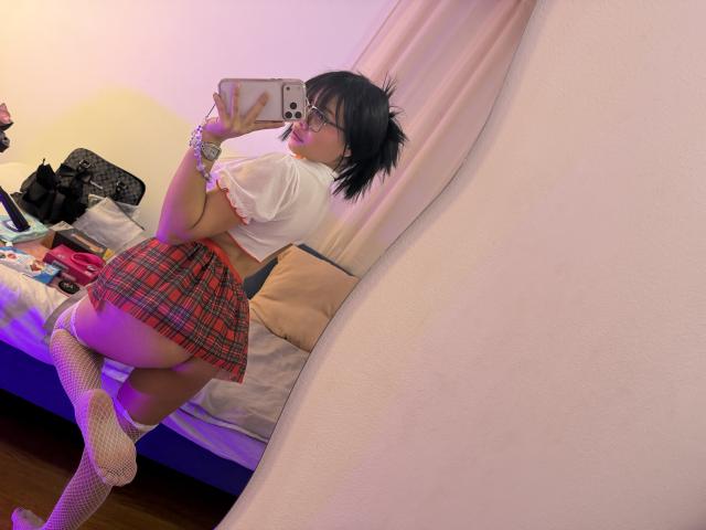VannesaSaenz with black hair, glasses, white top, red plaid skirt, fishnet socks, showing her ass, kneeling, taking a mirror selfie in a bedroom.