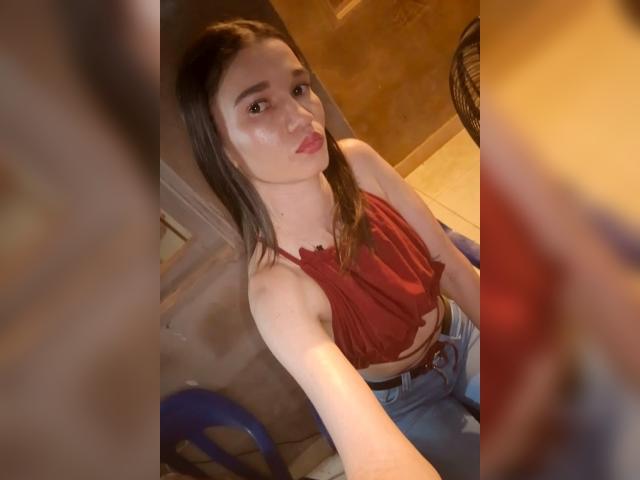 JennaDerose with pouty lips, brown hair, red halter top, showing small tits, and blue jeans, taking a selfie from above, in a dimly lit room.