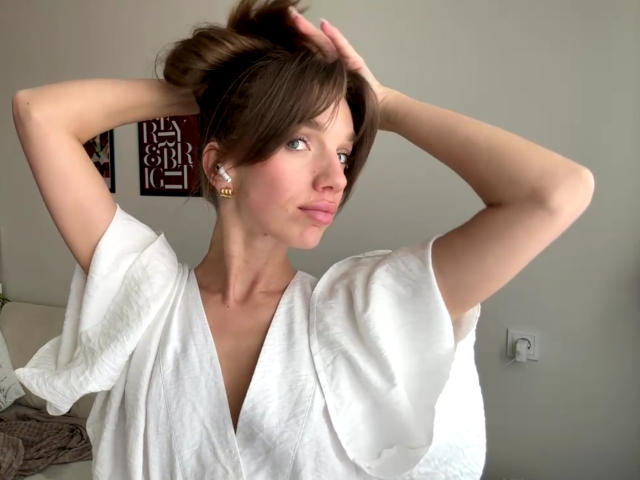 PortiaKomer with hands in brown hair, wearing a white V-neck shirt, small gold earrings, looking to the side with a slight smile, soft light on her face.