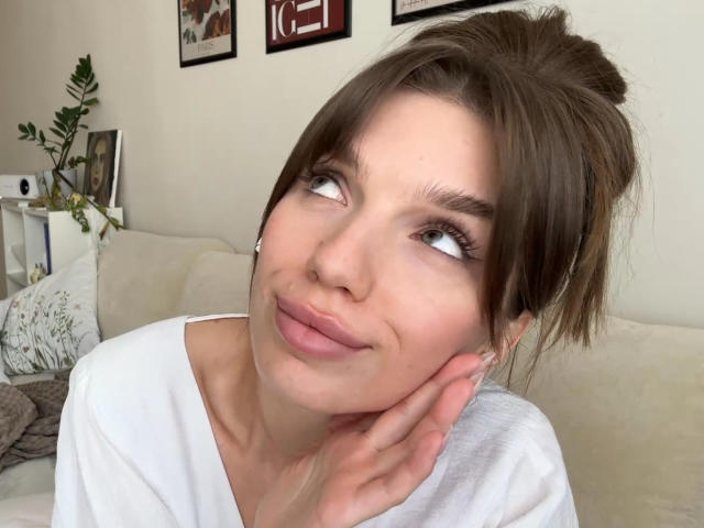 PortiaKomer with brown hair in a messy bun, white shirt, looking up with a cute, slightly smirking face, hand touching her cheek. Her tits are small, eyes are blue.