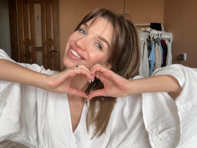 PortiaKomer, smiling, wearing a white bathrobe, makes a heart shape with her hands, brown hair in a ponytail, blue eyes, white walls in the background, door and clothes visible.