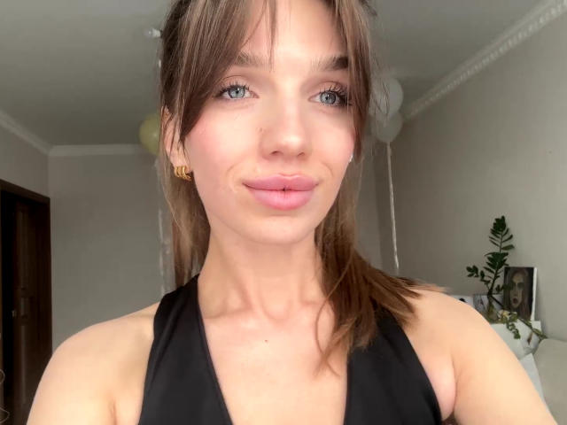 PortraitKomer has light blue eyes, pink lips, and brown hair in a ponytail, wearing a black halter top, with a subtle smile, and gold ear cuffs.