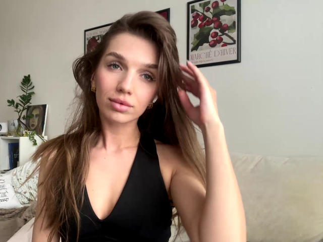 PortiaKomer with long brown hair, wearing a black halter top, has a slight smile, and is touching her hair with her right hand. Her tits are visible.
