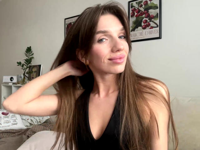 PortiaKomer has long brown hair, a cute face, and small tits, wearing a black halter top, smiling with a hand in her hair, in a bright room.
