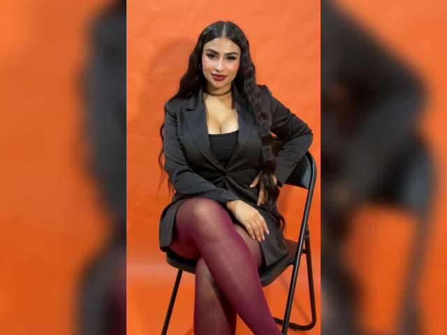 MiilaWx with long black hair, red lipstick, black blazer, black dress, and red tights, sitting on a black chair against an orange background, showing her boobs and crossed legs.