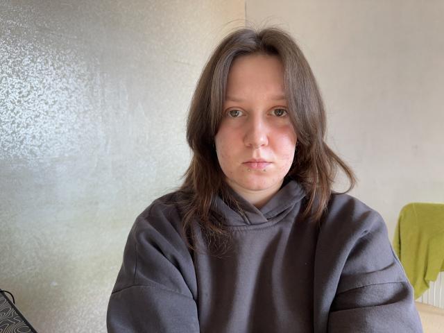 RubyOiler has pale skin, brown eyes, and medium-length brown hair. She's wearing a grey hoodie and has a small patch of greenish skin near her mouth.
