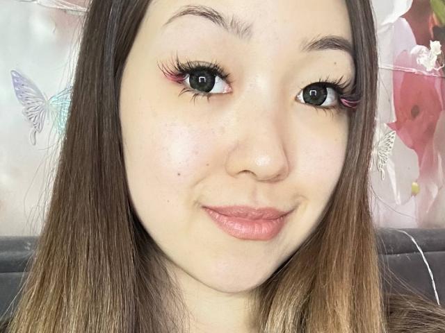 RenyLime has light skin, brown eyes with pink eyeliner, long eyelashes, and pink lips. She's smiling with straight brown hair, and small moles are on her left cheek.