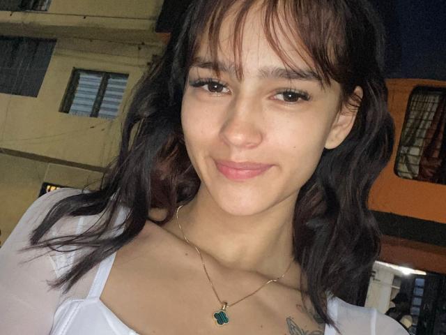 MelodyMansson has wavy brown hair, light skin, and a small smile. She's wearing a white top, a necklace with a green gem, and has a small tattoo on her chest.