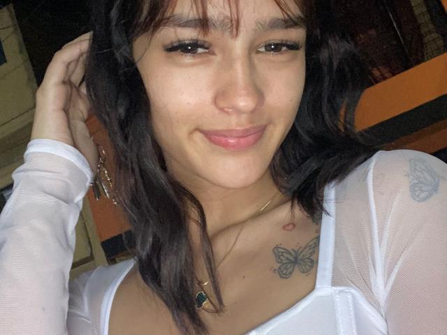 MelodyMansson has a cute smile, brown eyes, and wavy black hair. She's wearing a white sheer top, showing a butterfly tattoo on her right chest. Her tits are small.