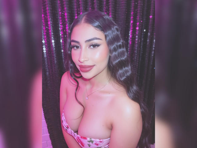 MiilaWx with wavy black hair, pink lipstick, and a floral strapless dress, showing her tits, smiling, against a shiny black sequined background. She's wearing a delicate necklace.
