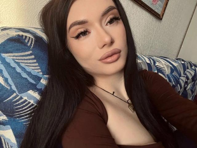 AlessandraShoe with long black hair, full lips, and thick eyelashes, wearing a brown top, showing her small tits, sitting on a blue patterned couch.