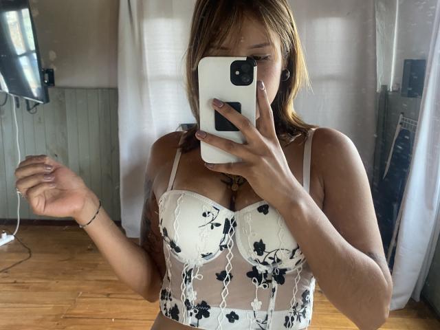 KiaraHernandezX with medium tits in a white floral lace bra, holding a phone in front of her face, standing in a room with wooden floors and white curtains.