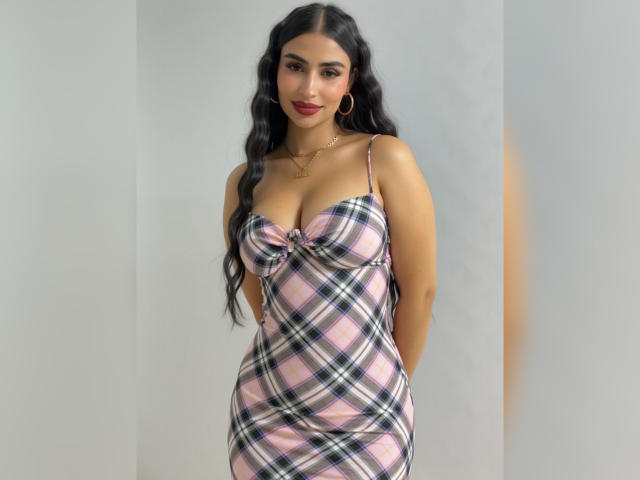 MiilaWx with long black hair, wearing a pink and blue plaid dress that shows her big boobs and small waist, standing with arms behind her back, smiling.
