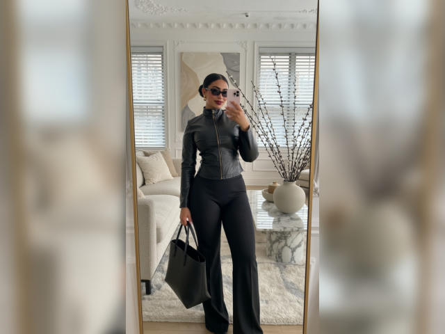 MiilaWx with black leather zip-up top and black flared pants, holding a black bag, standing in front of a mirror, wearing dark sunglasses, red lipstick, and black hair.