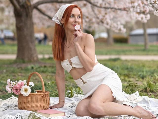 MiraGi with red hair, white tube top, and skirt, sitting on a lace blanket, holding a pink flower, wicker basket with flowers, books, and green grass.