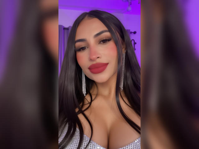 MiilaWx with long black hair, red lipstick, and sparkly earrings, showing off her big boobs in a silver, glittery top, looking into a mirror with purple lighting.