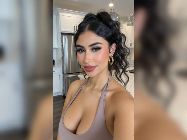 MiilaWx with wavy black hair in a high pony, wearing a tan halter top, showing her big boobs and pink lipstick, smiling in a kitchen with a blurred reflection.
