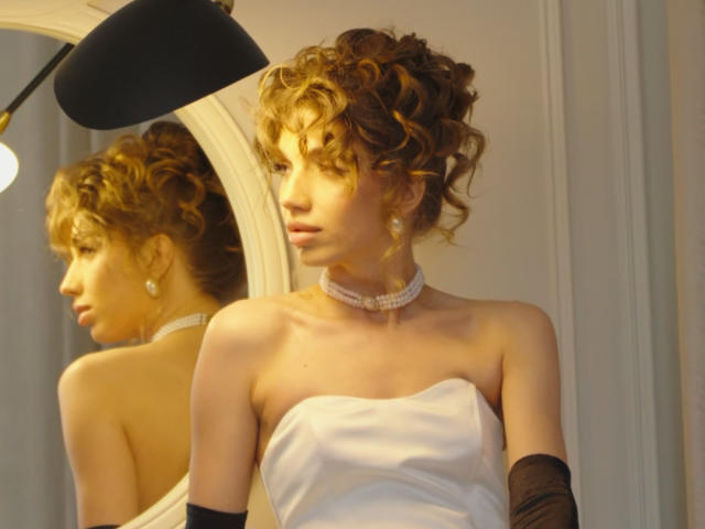 PortiaKomer with curly blonde hair, wearing a strapless white dress, black gloves, and a pearl choker, standing in front of a mirror reflecting her profile.