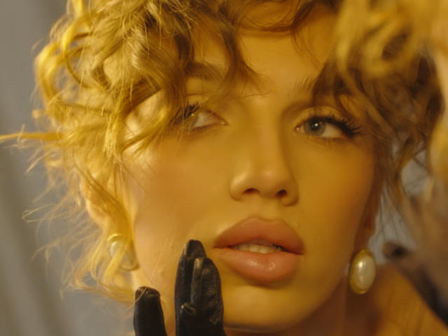 PortiaKomer's face with curly blonde hair, looking to the side, wearing black gloves, pink lips, and pearl earrings, in a shot with soft yellow lighting.