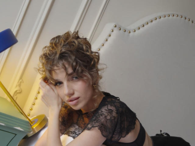 PortiaKomer with curly brown hair, wearing a black lace top, leans on a yellow desk, looking seductively at the camera, her tits partially visible, in a stylish room.