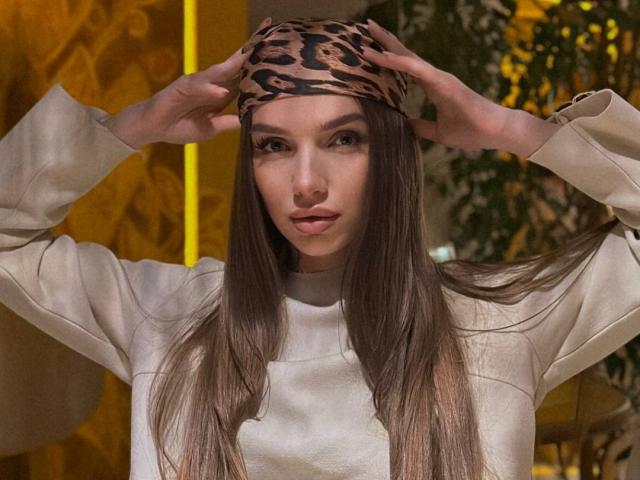 PortiaKomer with long brown hair, wearing a leopard print headscarf and beige puffy-sleeve top, hands adjusting headscarf, looking straight at camera. She has a nose ring.