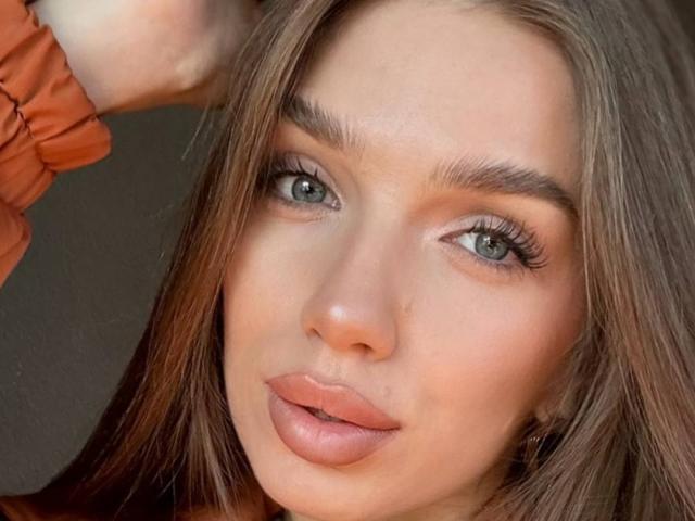 PortiaKomer's face, with her green eyes, full pink lips, and brown hair, wearing an orange shirt, has her hand up, showing off her eyebrows and long lashes.