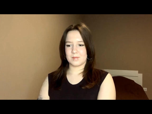 RubyOiler with pale skin, brown hair, small tits, black sleeveless top, hoop earrings, subtle tattoo on left arm, slight smile, brown wall, bed in background, soft lighting.